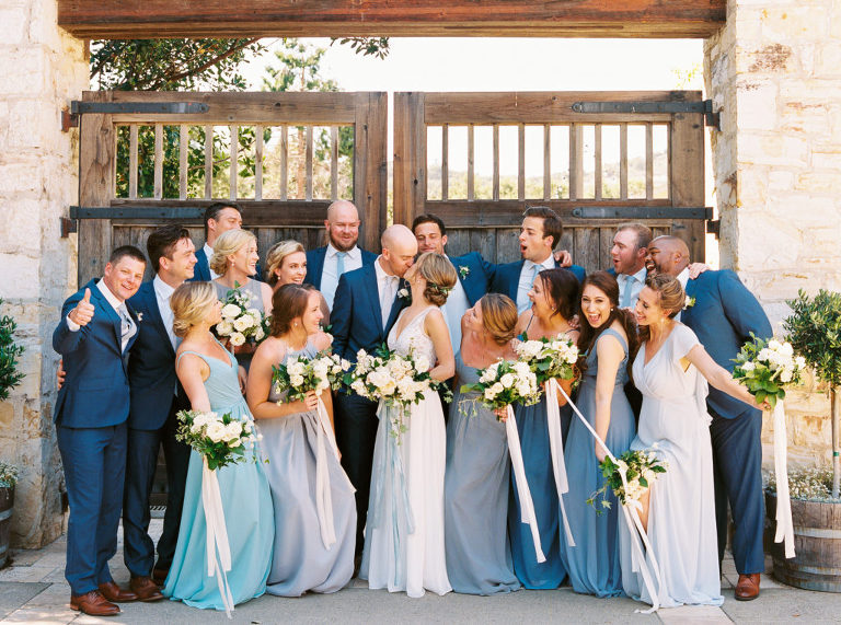 Large bridal party celebrating with newlyweds in front of large rustic gates at Holman Ranch