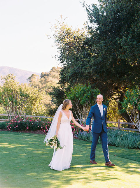Newlyweds walking hand in hand through the lawn area at Holman Ranch