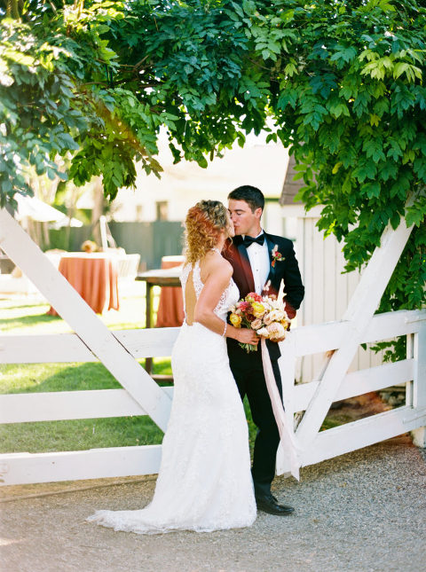 Newlyweds kissing in front of overgrown greenery and white gate in Los Olivos