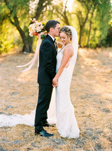 A sun filled photo of a bride and groom holding each other. The bride has a large colorful bouquet in her hand. 